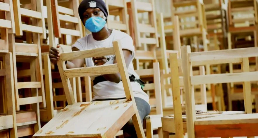 A young African woman holds an unvarnished chair, with other unfinished chairs around her.