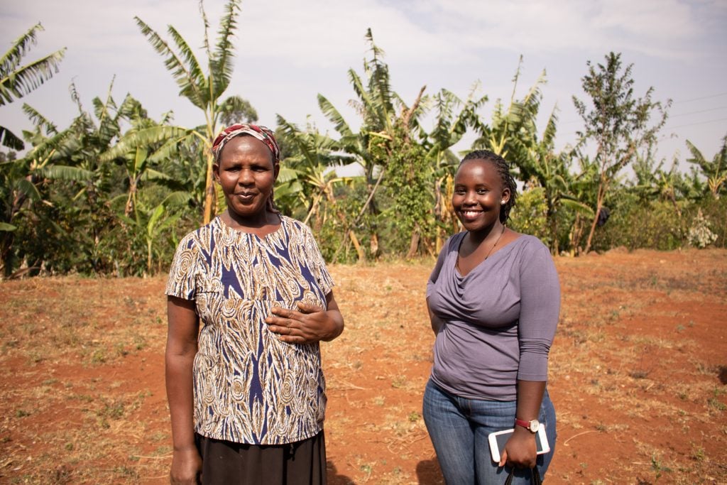 Two Ugandan women stand in front of coffee trees.
