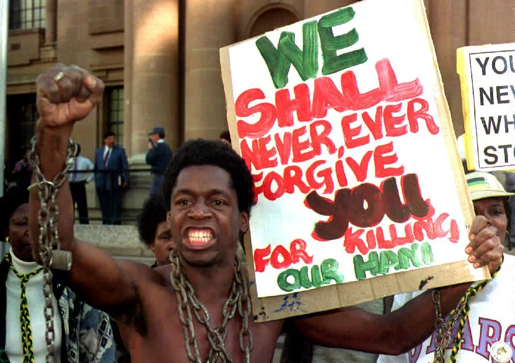 An African man wearing chains and carrying a placard saying "We will never, ever forgive you for killing our Hani"